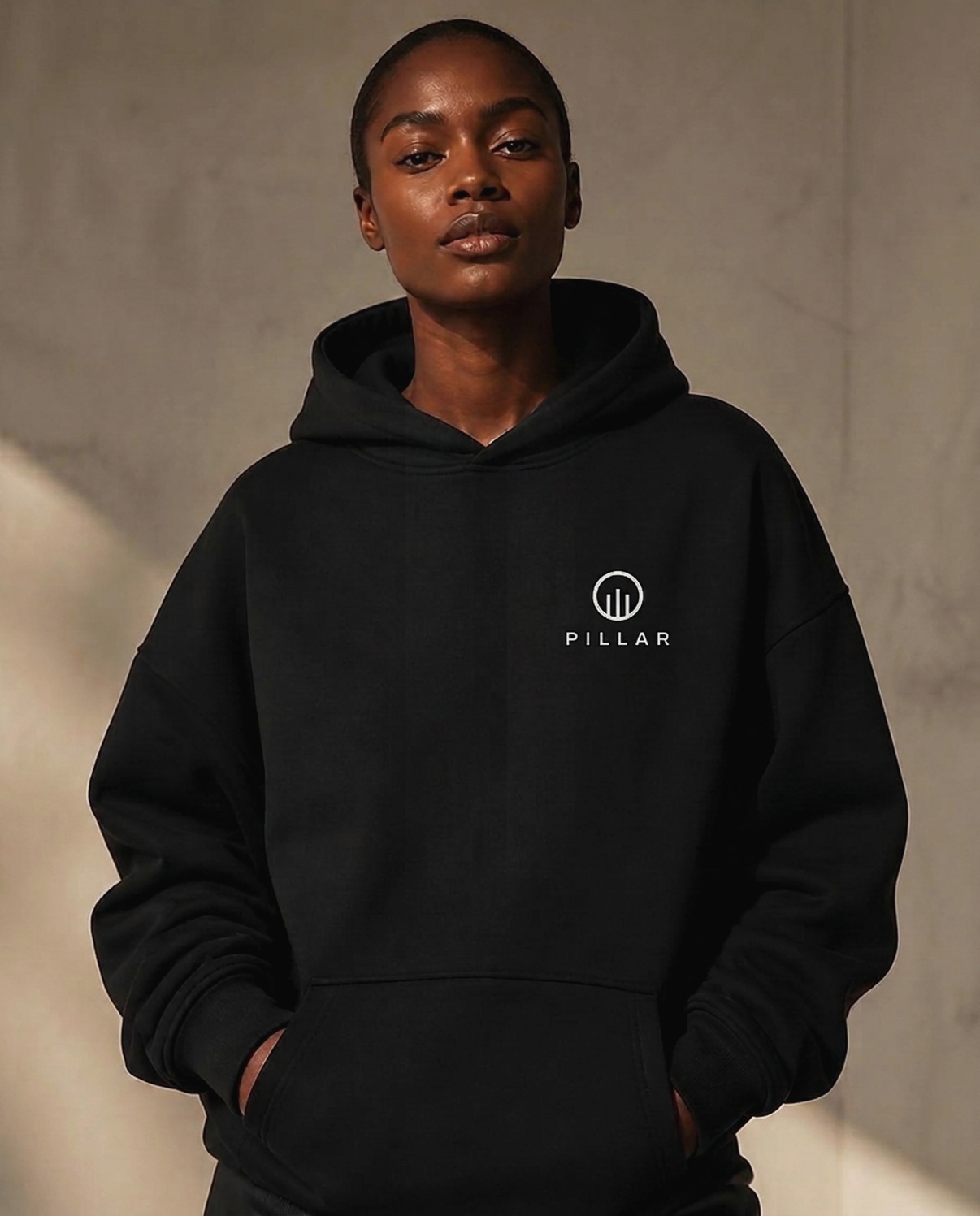 Black female model wearing the black PILLAR Forged Hoodie with the embroidered left-chest lockup